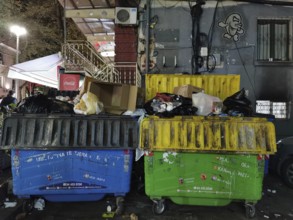 Two colored, overcrowded dumpsters on a street at night with graffiti on the wall, Himare, Albania