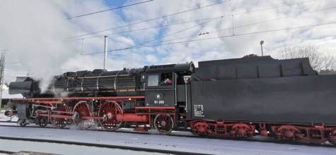 A black, historic steam locomotive emits smoke and stands on snow-covered tracks in winter,