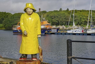 Statue of a fisherman in yellow raincoat at harbour in front of anchored boats with green hills in