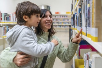 Woman and son smiling together at bookshelves, choosing a book in a library, bonding over reading