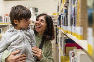Mother holding her young child, both smiling and interacting happily while standing by shelves