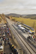 Numerous people gather at a train station in a rural area with rolling hills in the background,