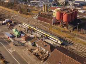 Industrial site with a passing train, surrounded by people and various buildings, opening of the