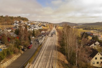 A railway line runs through a village with houses and trees in a hilly winter landscape, opening of