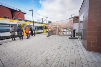 People wait on the railroad track next to a yellow train, a large poster is visible, battery