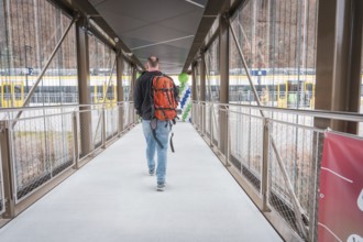 Man with backpack walking across bridge, yellow train in background, battery electric train Siemens