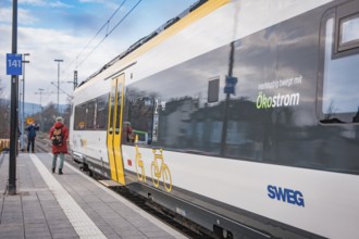A SWEG train with green energy branding stands at the station while a person with a backpack passes