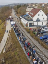 People wait for a train at a station along the tracks next to a road, opening of the Hermann Hesse