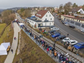 A crowd of people waits in a village along the railroad tracks next to a road, opening of the