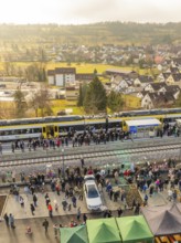 A train with people waiting on a platform in rural surroundings, opening of the Hermann Hesse