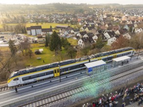 A train is standing at a train station, people are standing to the right, a village and fields in