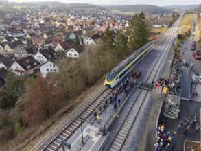 A train stops at a rural train station with surrounding people. The surrounding area shows a