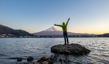 Young man stretching his arms in the air, standing on rocks in Lake Kawaguchi, view of Mount Fuji