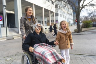 Three generations walking outdoors, smiling grandmother in wheelchair pushed by her daughter while