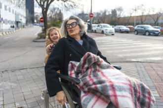 Happy grandchild pushing a senior woman with grey hair and glasses in a wheelchair, enjoying an
