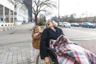 Granddaughter pushing her joyful grandmother in a wheelchair during an outdoor walk on a sunny day,
