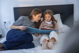 Mother and young daughter share a quiet bedtime moment reading together in bed, fostering