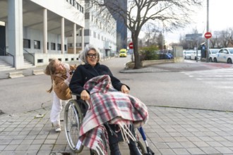 Granddaughter joyfully pushing her smiling grandmother in a wheelchair, exploring the city together