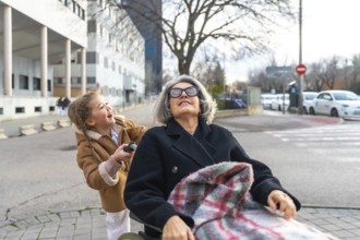 Happy granddaughter pushing senior grandmother in a wheelchair, enjoying a sunny day outdoors and
