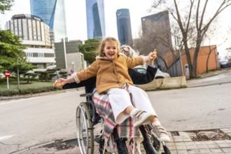 Happy little girl laughing and spreading arms, sitting on an older woman's lap in a wheelchair,
