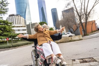 Happy granddaughter sitting on grandparent's lap in a wheelchair, holding hands and laughing with