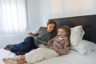 Mother and young daughter bonding over a book in bed, sharing a laugh and enjoying quality time