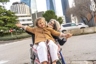 Smiling granddaughter sitting on grandmothers lap in a wheelchair as they enjoy an outdoor stroll