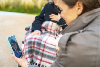 Daughter visiting grandmother in a wheelchair outdoors, sharing a video call with another family