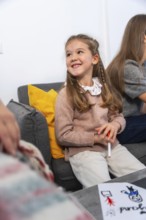 Happy little girl sitting on a sofa, smiling while holding a marker and showing a homemade drawing