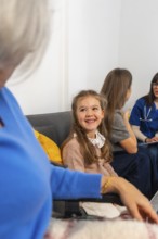 Smiling young girl visits grandmother in a wheelchair at a nursing home, surrounded by family and a