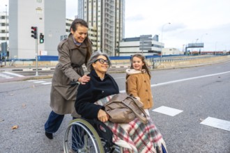 Family members assisting senior woman in wheelchair outdoors, granddaughter walking alongside while