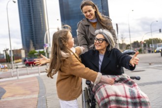 Smiling grandmother in wheelchair reaching to embrace granddaughter while adult woman pushes them