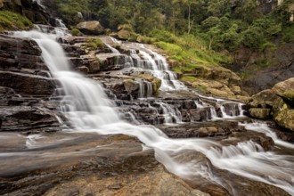 An impressive waterfall flows over stepped rocks surrounded by green nature and thick trees,