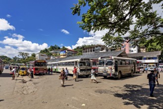 Busy bus stop with people and buses under a sunny sky with green trees, Kandy bus station in Sri
