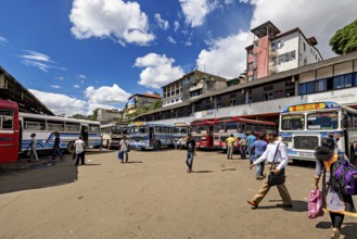 People walk past a bus stop surrounded by various buses and clouds in the sky, Kandy bus station in