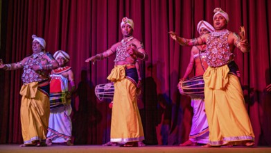 Men in traditional clothing play drums on a stage in front of a red curtain, folklore and dancers