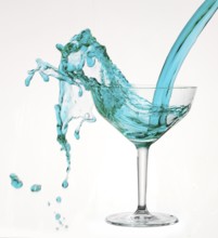 A powerful flood of blue liquid pours into a cocktail glass and spills over, studio shot against a