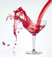 A powerful flood of red liquid pours into a cocktail glass and spills over, studio shot against a