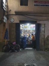 Small shop at night with open gate and bicycle, passers-by on the street in the Medina, Marrakech,