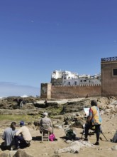 Painters paint the city view on the coast in front of old city walls and rocks under a blue sky,