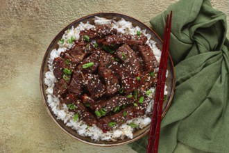 Crispy beef, with green onions and sesame seeds, on a rice pad, in a bowl with chopsticks, on a