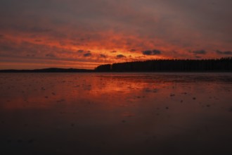A dramatic sunset over a still, frozen lake and forest with orange-red hues in a cloudy sky,