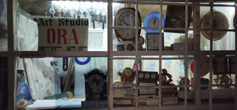 A shop window of an art workshop with various clocks and a sign, illuminated at night, Berat,