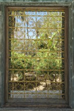 A richly decorated iron window provides a view of a green garden under a clear sky