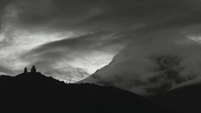 Dramatic silhouette of church buildings in front of a cloud-covered mountain, Kazbegi,