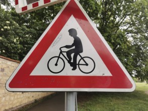 Road sign for bicycles, red and white triangle in the foreground, trees in the background, Czech