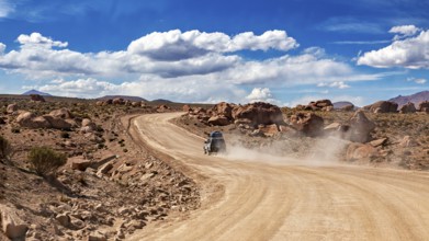 A vehicle drives on a dusty desert road through rocky landscapes under blue skies, The landscape of