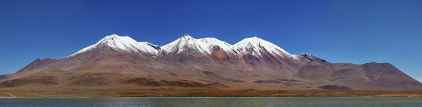 Snow-capped mountains above a desert plateau are reflected in a calm lake under a clear blue sky,