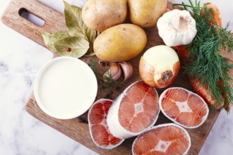 Potatoes, salmon, garlic, onion, and herbs are placed on a wooden cutting board. These ingredients