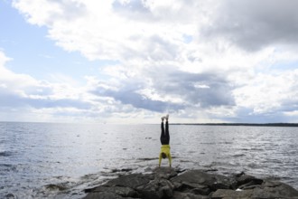 Young woman girl doing a handstand on rocks at Lake Vänern under cloudy sky, Värmlands län, Sweden